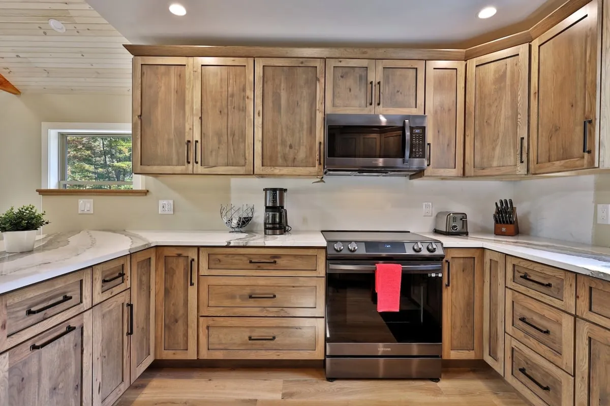 A spacious, modern kitchen with wooden cabinetry, stainless steel appliances, marble countertop, and a red dish towel. The area is warmly lit and inviting, ideal for group cooking or gathering over shared meals.