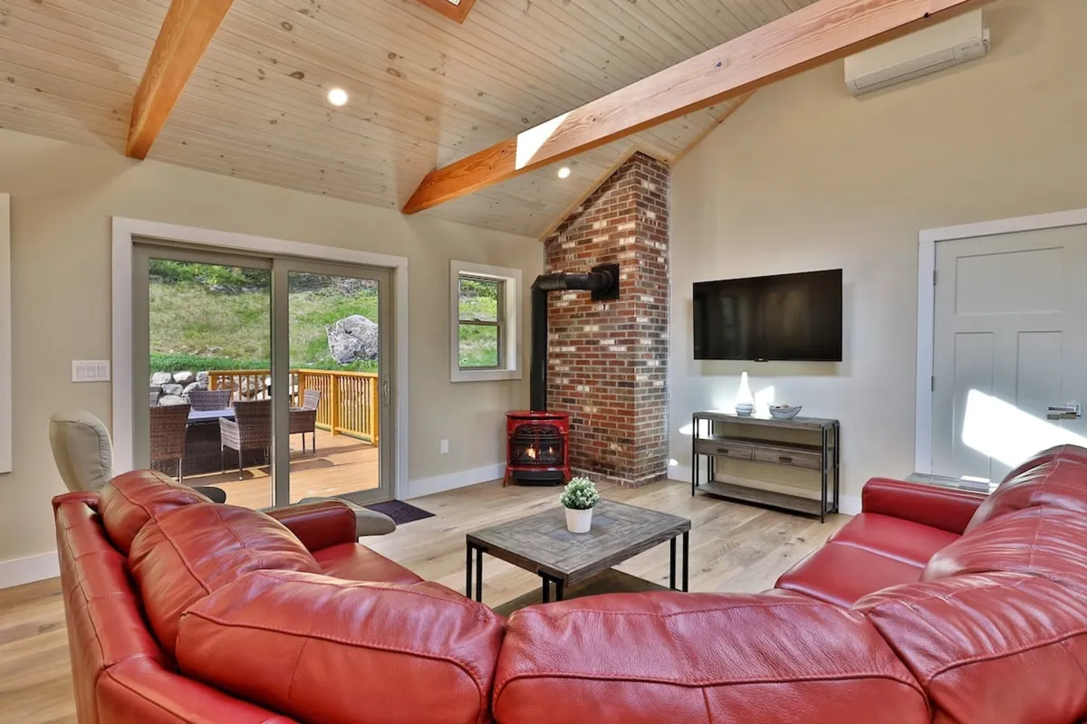 A spacious living area featuring a red sectional sofa, a classic brick fireplace, and large windows overlooking a wooden deck. The arrangement is warm and comfortable, inviting relaxation after a day outdoors.