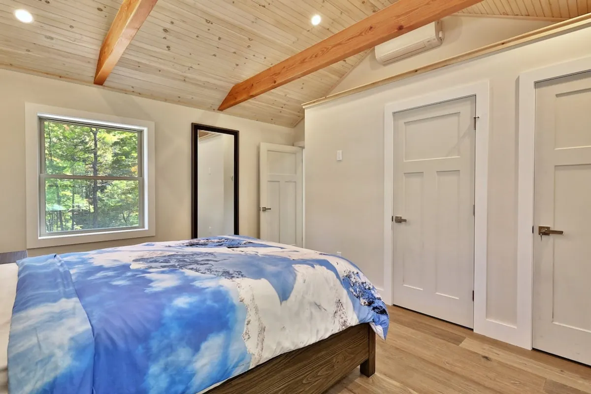 A bright, airy bedroom with blue and white bedding, a large window letting in natural light, and exposed wooden beams. The view of greenery outside adds to the calming, restorative feel.