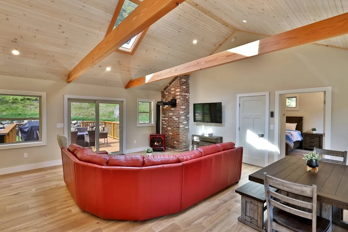 A cozy and welcoming living/dining area with a red sectional sofa, a brick fireplace, a wooden dining table, and expansive windows showcasing serene views outside. The arrangement invites group gatherings, relaxation, and socializing after a day of exploring.