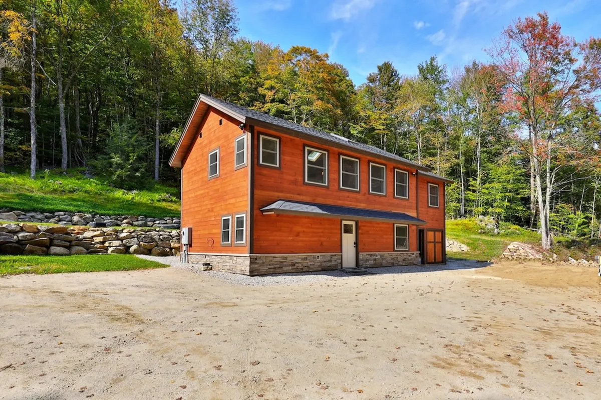 The image shows the inviting wooden exterior of Peak Hill Home, nestled among tall trees in a quiet, forested setting. Large windows reflect the natural surroundings, and a spacious gravel driveway leads up to the house. The setting evokes tranquility, with the lush Vermont woods enveloping the property and hints of a large deck for outdoor gatherings.