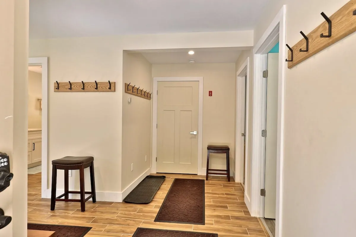 A close-up of wooden coat hooks mounted on the wall, a stool, and a doorway leading further into the home. The scene captures the details and practical elements of the entry space, emphasizing first impressions and ease of access.