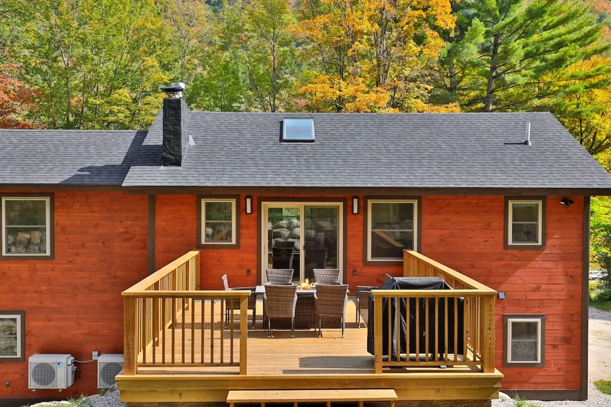 The image presents a wooden deck with outdoor furniture, bordered by large windows and framed by vibrant autumn foliage. The cozy arrangement suggests spaces for morning coffee, outdoor dining, or simply soaking in the serene woodland setting.