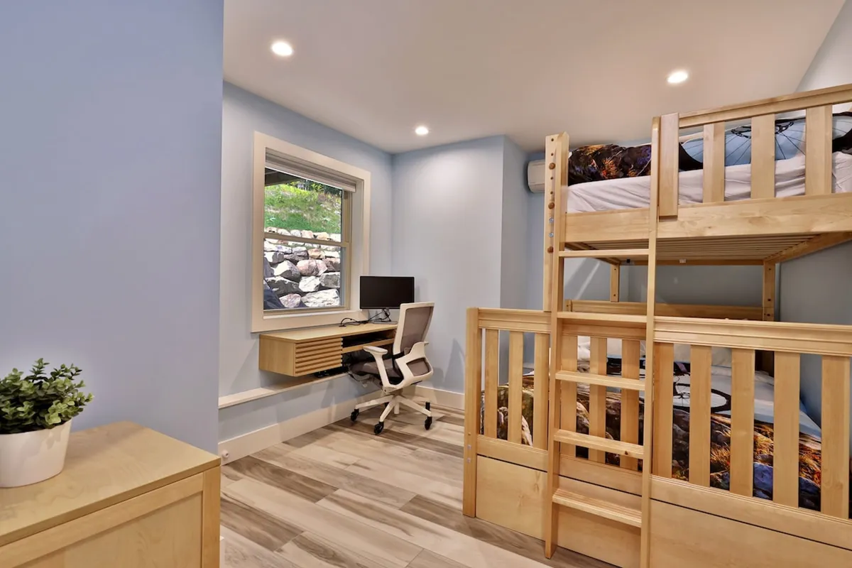 A cozy workspace is set up next to a window, allowing abundant natural light to flood the room. The bunk bed nearby offers a comfortable spot to relax or nap, while the desk features a computer setup perfect for remote work or study. The window view hints at the peaceful, natural surroundings outside, enhancing the atmosphere of productivity and calm.
