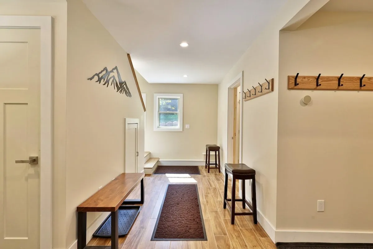 The image shows a cozy mudroom-like area with a wooden bench, mountain wall art, stools, and several coat hooks. The layout is ideal for guests to sit and remove boots or hang outdoor clothing, emphasizing the transition from outside to inside.