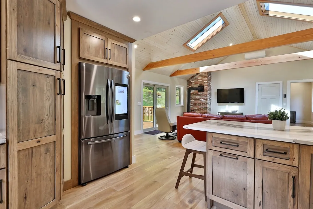 A spacious, inviting kitchen and living area featuring a large island with bar stools, warm wooden cabinetry, a stainless steel refrigerator, and a cozy brick fireplace. Large windows allow natural light to stream in, and subtle glimpses of greenery outside hint at the home's wooded setting.