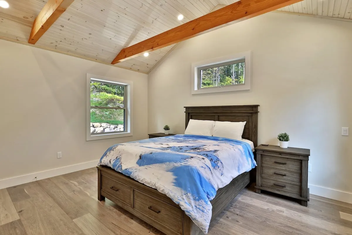 A serene bedroom featuring a bed with blue and white bedding, a wooden nightstand, and a window showcasing lush greenery outside. The ambiance is peaceful and perfect for relaxation after an event.