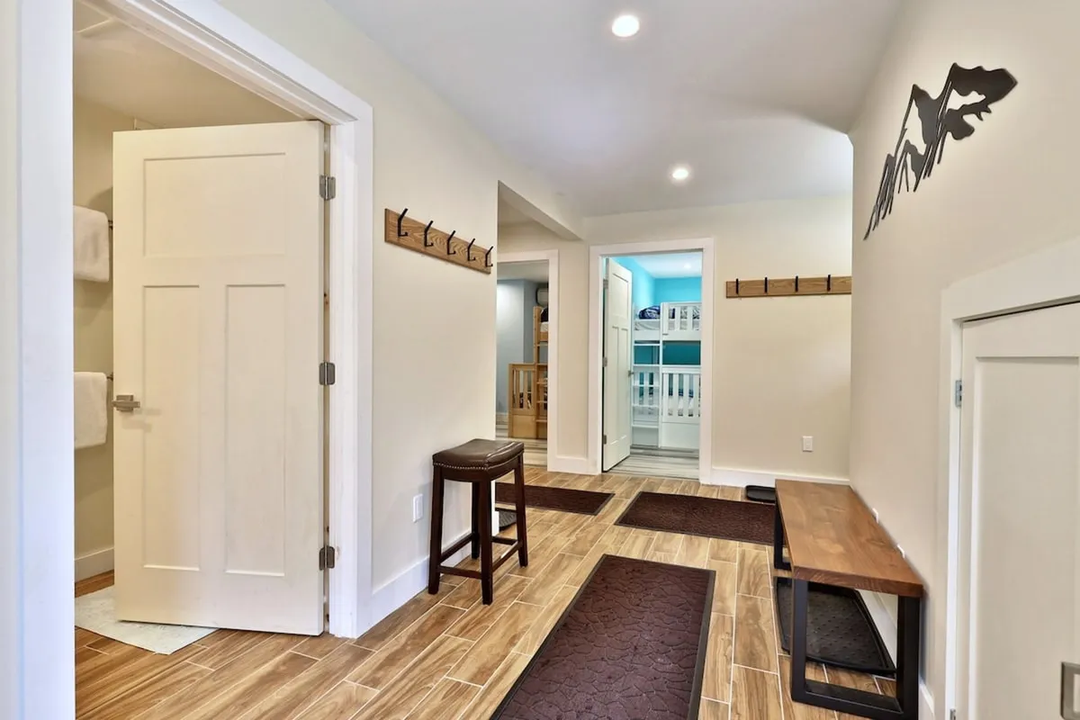 This image captures a functional and inviting entryway featuring a bench, coat hooks for gear storage, an open door to another room, and a wall-mounted mountain decoration. The space is well-lit and organized, giving guests a sense of arrival and convenience for storing outdoor equipment.