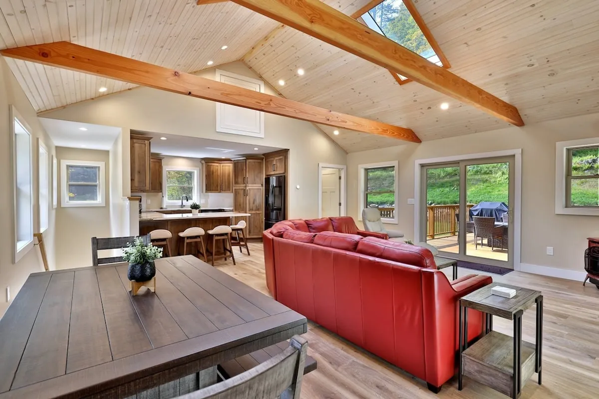 A welcoming interior with a red sofa, dining table, large windows allowing natural light to flood in, and a kitchen island in view. The space feels designed for family meals, celebrations, and relaxed mornings with coffee.