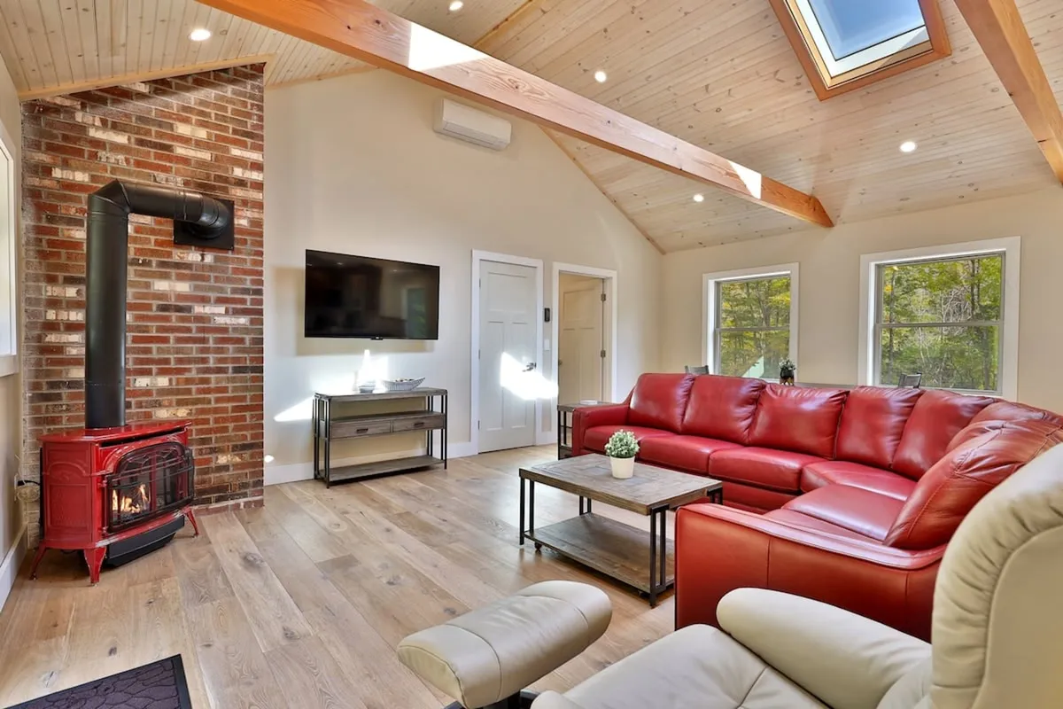 A snug living area with a red leather sofa, rustic wooden beams, a brick wall featuring a fireplace, and a television. The setup invites relaxation and intimate gatherings, ideal for unwinding after an energetic evening.
