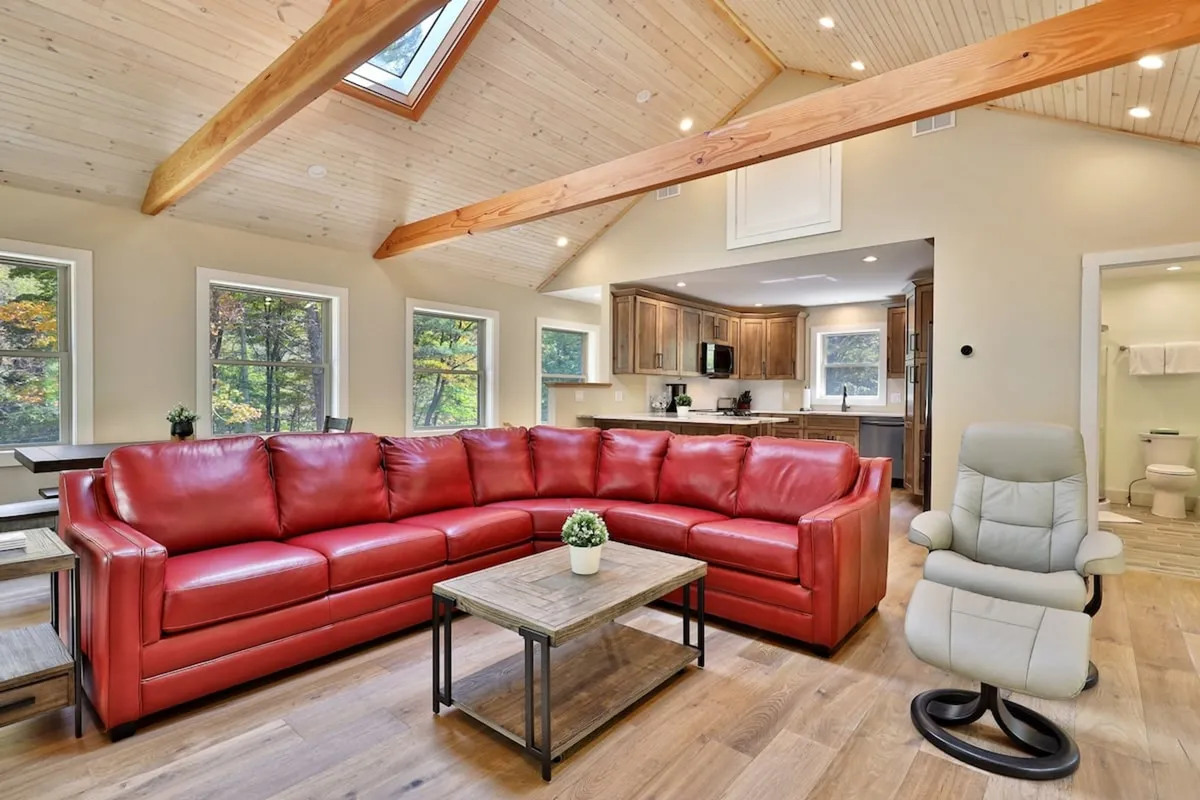 A wide-angle view of the open-plan living area showcasing a plush red sectional sofa, a wooden coffee table, large windows letting in abundant natural light, and the adjacent kitchen area. The layout highlights vaulted ceilings and the seamless flow between living, kitchen, and dining spaces, emphasizing togetherness and comfort.