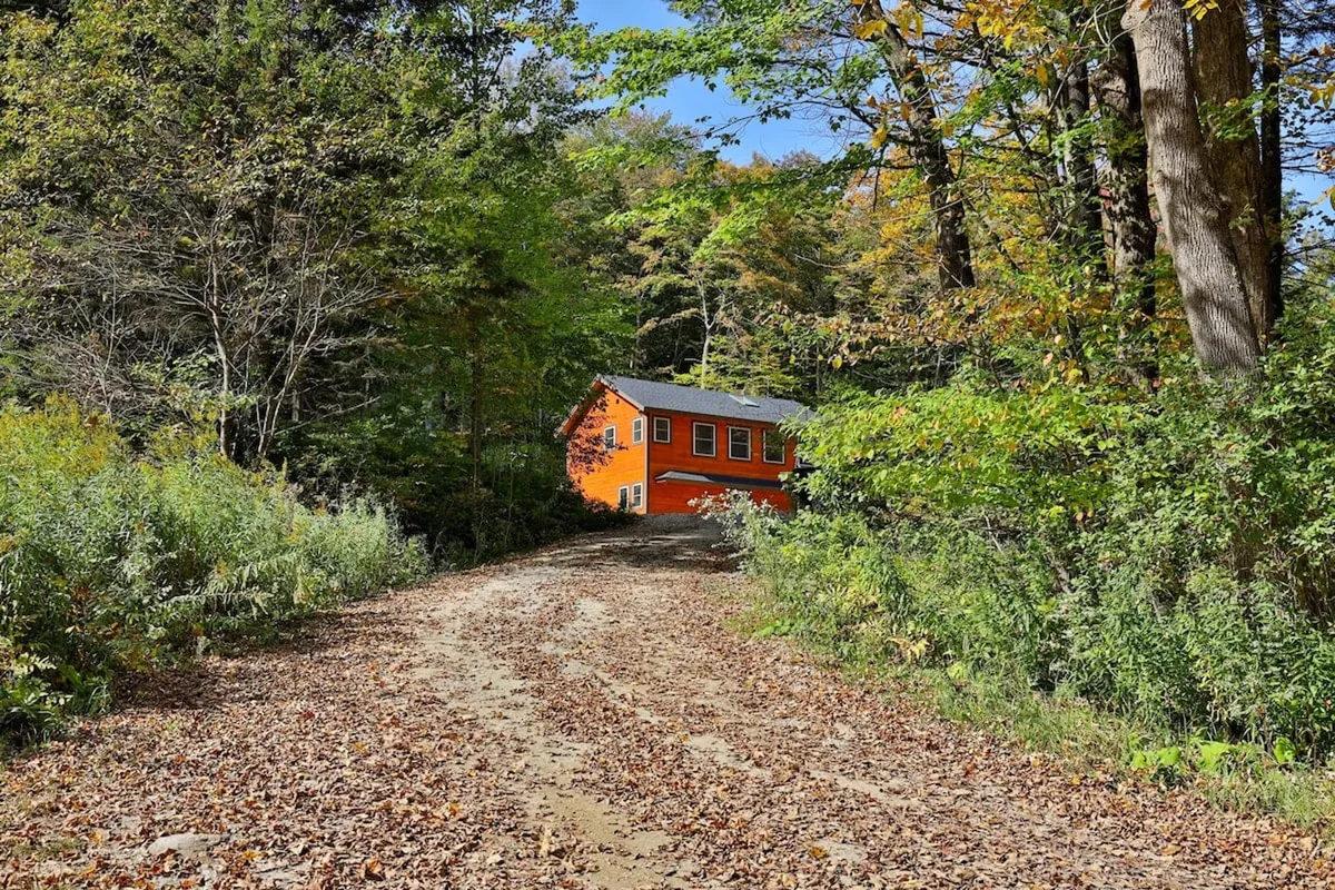 An inviting exterior view of the orange Peak Hill Home surrounded by trees, with a leaf-covered path leading to the entrance. The scene conveys a sense of privacy and natural beauty, situating the home within Vermont’s scenic landscape.